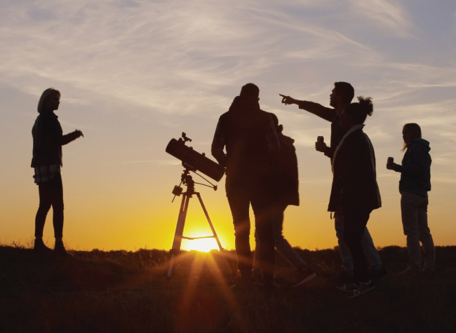 Group of people surrounding a telescope practicing astronomy, with a sunset backdrop